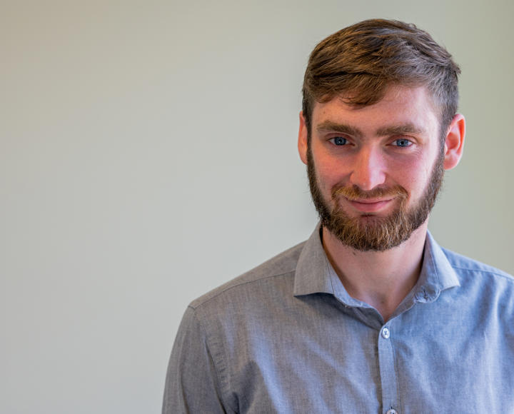 A young male graduate trainee. He is wearing a light grey shirt and has brown hair and facial hair, with blue eyes. 